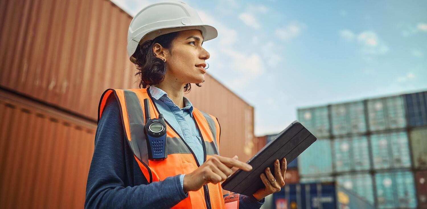 Eine Frau mit Warnweste, Helm und Tablet in der Hand auf einem Gelände auf dem Container stehen.