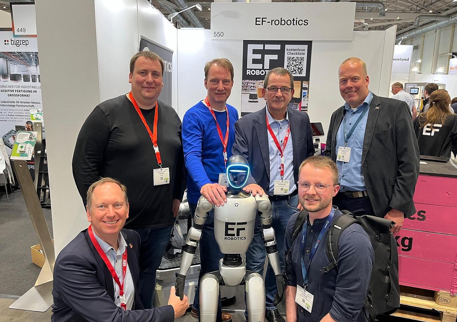 Eine Personengruppe auf einem Messestand. In der Mitte steht ein Roboter.
