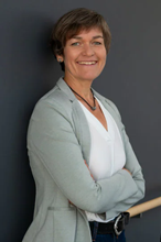 Foto einer Frau mit kürzeren dunklen Jahren die an einer Wand gelehnt steht und einen Anzug trägt. Sie lächelt in die Kamera.