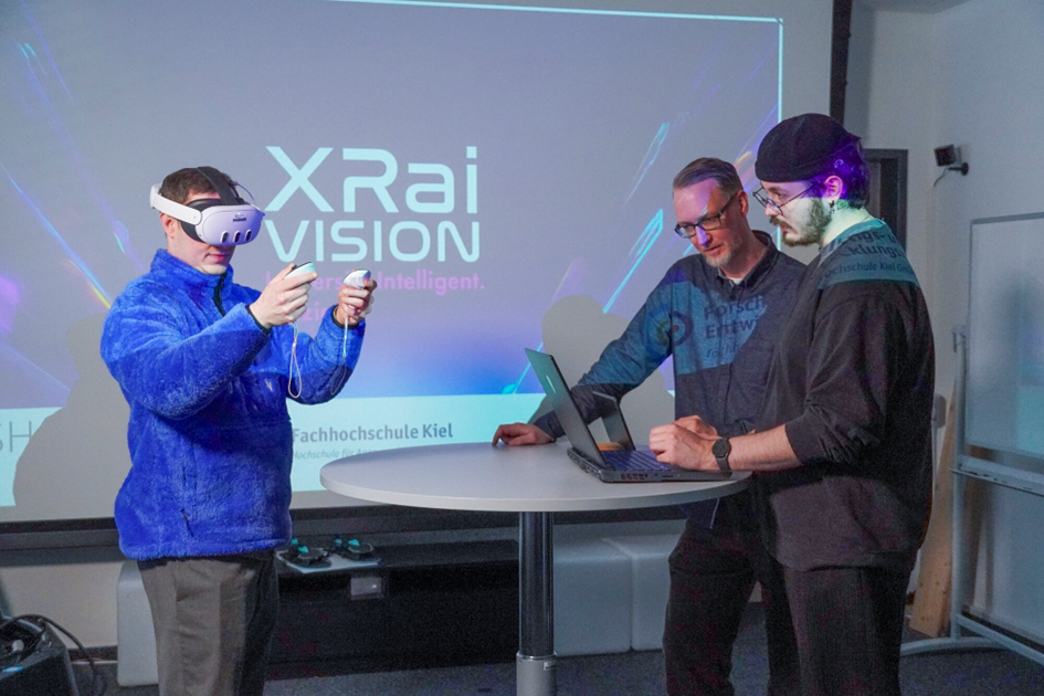 Ein Mann mit einer VR-Brille auf, steht vor einem Stehtisch an dem zwei weitere Männer stehen.