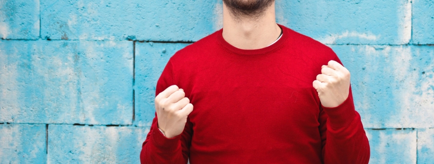 Ein lächelnder Mann in einem roten Pullover steht vor einer hellblauen Wand und zeigt Freude mit erhobenen, geballten Fäusten.