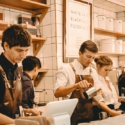 Schnappschuss von fünf Caféangestellten hinterm Tresen. Es scheint viel los zu sein, jeder ist beschäftigt: Einer bedient die Kasse, ein anderer gießt Milchschaum in die Tasse. Der Hintergrund ist weiß gekachelt, mit Holzregalen, auf denen Produkte ausgestellt sind.