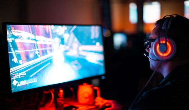 Im Vordergrund: ein Mann, der am PC ein Spiel spielt. Er trägt Headsets, auf dem Bildschirm sieht man das Spiel. Das Zimmer im Hintergrund ist verdunkelt.