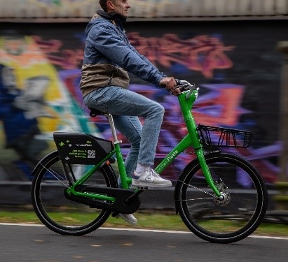 Ein seitlicher Blick auf einen Radfahrer, der mit einem grünen Fahrrad der Kieler Sprottenflotte vor einer mit Graffiti besprühten Betonmauer fährt. Der Radfahrer trägt eine Jeanshose und eine Jacke.