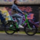 Ein seitlicher Blick auf einen Radfahrer, der mit einem grünen Fahrrad der Kieler Sprottenflotte vor einer mit Graffiti besprühten Betonmauer fährt. Der Radfahrer trägt eine Jeanshose und eine Jacke.