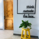 links eine Holztür, rechts ein gelber Hocker mit einer Pflanze. Darüber hängt ein schwarzer Rahmen mit dem Spruch: think outside the box