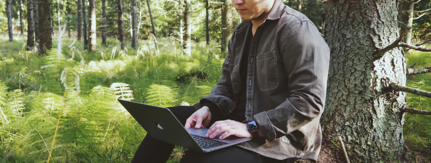 Mann sitzt mit Laptop auf den Beinen, angelehnt an einem Baum inmitten eines Waldes.