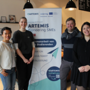 Auf dem Bild stehen drei Personen in einem Café, die in die Kamera schauen. In der Mitte steht ein Werbebanner mit der Aufschrift Artemis.