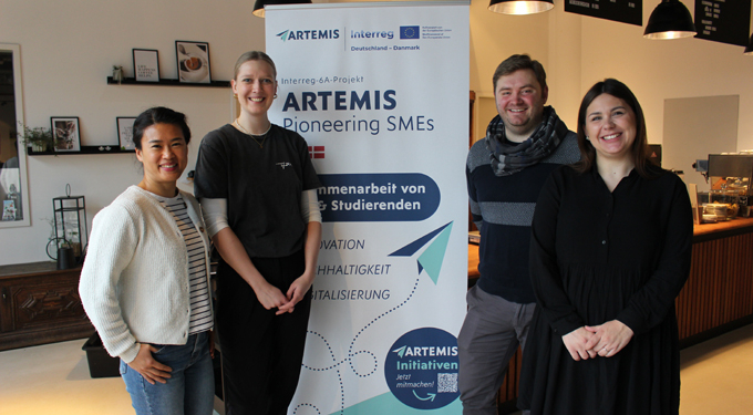 Auf dem Bild stehen drei Personen in einem Café, die in die Kamera schauen. In der Mitte steht ein Werbebanner mit der Aufschrift Artemis.