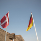 Eine dänische und eine deutsche Flagge an Fahnenstangen vor einem Reetdachhaus.