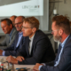 Auf dem Foto sind vier Männer in Anzügen bei einer Pressekonferenz zu sehen, die im Gespräch vertieft sind.