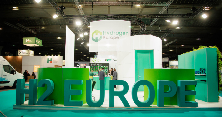 Das Bild zeigt eine Messehalle und einen Messestand vor dem in großen Buchstaben steht H2 Europe.