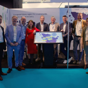 Eine Gruppe von Personen auf einem Messestand von Clean Energy Valley. In der Mitte ein Bildschirm auf dem eine Karte von Schleswig-Holstein gezeigt wird. Alle schauen in die Kamera und lächeln.